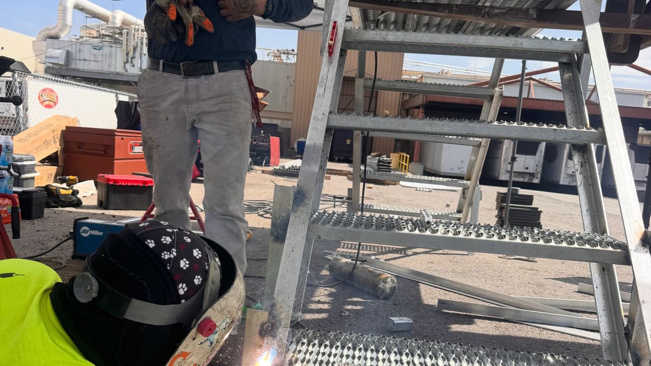 A welder in a Pro-fusion shirt welding the rail of a galvanized stair on a rooftop, under a canopy.