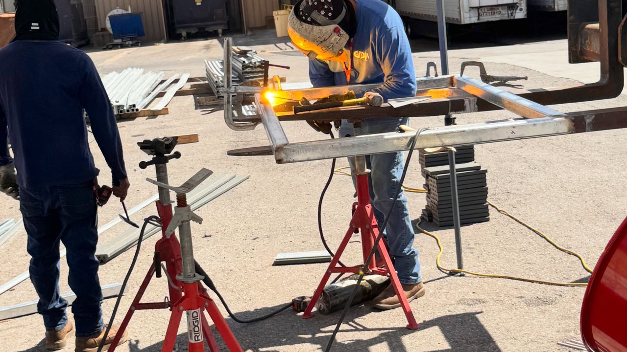 A welder under a canopy welding a frame on pipe stands, with a second build in progress behind him.