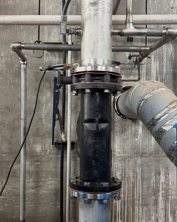 A black flanged valve fitting on a vertical stainless pipe against a concrete wall.