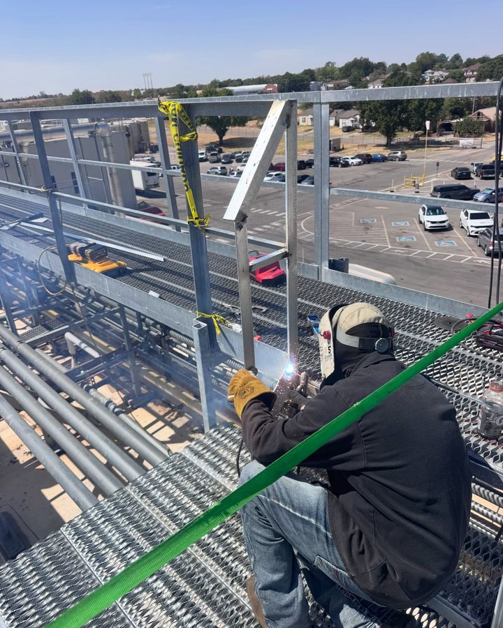 A welder sitting on galvanized grating, welding a handrail post on a rooftop catwalk above a parking lot.