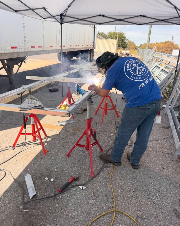 A welder in a blue Pro-fusion shirt welding an aluminum frame on red pipe stands, sparks off the joint.